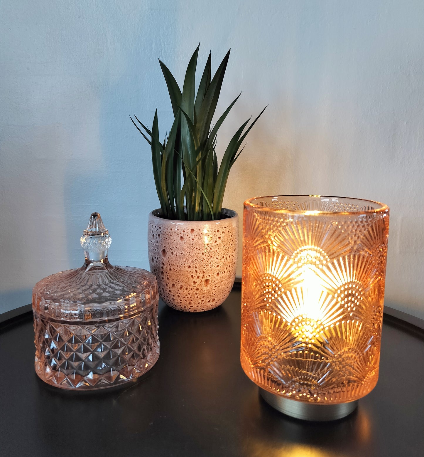 Led bordlampe rosa farvet med vifte møster med lys i. Lille glaskrukke i rosa farve. Lille kunstig bredbladet plante.